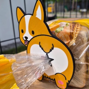 May include: A wooden corgi-shaped bread clip in orange, white, and black. The clip is designed to seal a bag of bread. The corgi has its tongue sticking out and is holding the bag closed. A loaf of bread is visible in the background.