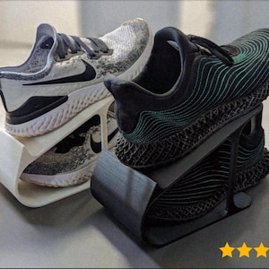 Könnte beinhalten: Zwei Paar Sportschuhe, die auf Schuhständern präsentiert werden. Ein Paar ist weiß und grau mit einem schwarzen Swoosh, das andere ist schwarz mit türkisfarbenen Akzenten. Die Schuhe stehen auf geschwungenen, minimalistischen Ständern.