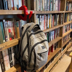 May include: A gray backpack with black trim hangs from a red hook on a wooden bookshelf filled with books. The books have various colored spines and titles. The bookshelf is made of light-colored wood. The image includes the text "@PieSupplies".