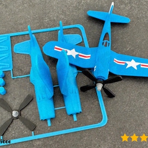 Op de afbeelding: Een blauwe plastic modelvliegtuigkit met een propeller en vleugels, gedeeltelijk gemonteerd. Het vliegtuig heeft witte sterren en rode strepen. De kit bevat ongemonteerde onderdelen op een blauw plastic frame.