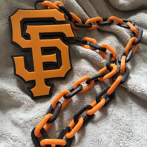 May include: An orange and black necklace featuring the San Francisco Giants logo. The pendant is a bold orange with a black outline, and the chain alternates between orange and black links. The necklace is likely made of plastic or a similar material.