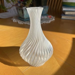 Puede incluir: Un jarrón blanco impreso en 3D con un diseño de remolino y textura. El jarrón tiene un cuello estrecho y una base ancha y redondeada.