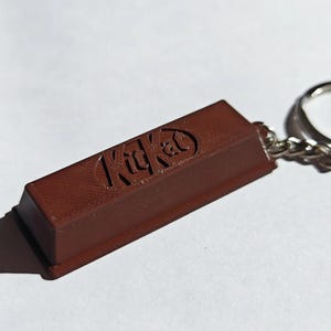 Może przedstawiać: Brelok w kształcie brązowego batona KitKat z wytłoczonym logo KitKat z przodu. Brelok jest przymocowany do srebrnego metalowego pierścienia i łańcuszka. Baton czekoladowy jest w jednolitym kolorze i wydaje się być wykonany z tworzywa sztucznego.