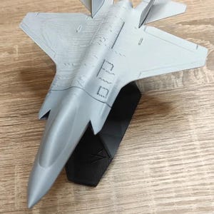 May include: A gray model airplane, resembling a fighter jet, is displayed on a black stand. The model has detailed wings, tail fins, and a pointed nose. The airplane is made of a light gray material.