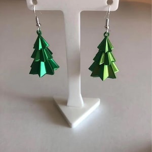 Puede incluir: Un par de pendientes verdes con forma de árbol de Navidad. Los pendientes están hechos de metal brillante y plegado y tienen un diseño en capas. Cuelgan de ganchos plateados. Los pendientes se exhiben en un soporte blanco sobre un fondo neutro.