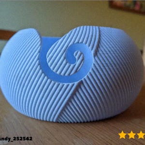 May include: A light blue yarn bowl with a spiral design. The bowl has a textured surface with vertical lines and a curved shape. The bowl is sitting on a wooden surface. The text "@MegaAndy_252542" is visible.