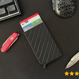 Puede incluir: Cartera negra con líneas diagonales, que contiene varias tarjetas de crédito. Una navaja suiza roja y un bolígrafo plateado también están sobre la superficie negra. Un ratón y un teclado de ordenador negros están en el fondo.