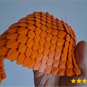 Może przedstawiać: Pomarańczowy, przypominający łuski przedmiot, prawdopodobnie element kostiumu lub przedmiot dekoracyjny, trzymany w dłoni. Obiekt ma teksturowaną powierzchnię i zakrzywiony kształt, z przedłużeniem w kształcie ogona. Tło jest jasnoniebieskie.