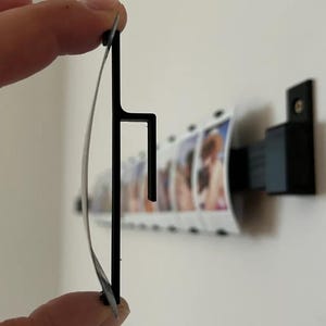 May include: A black metal photo display holder with a curved design. The holder is designed to hold a strip of photos.