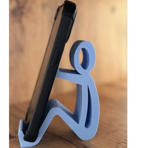 Könnte beinhalten: Ein hellblauer Telefonständer in Form einer sitzenden Person, die ein schwarzes Smartphone hält. Der Ständer besteht aus einem festen Material und ist so konzipiert, dass er ein Telefon aufrecht hält.