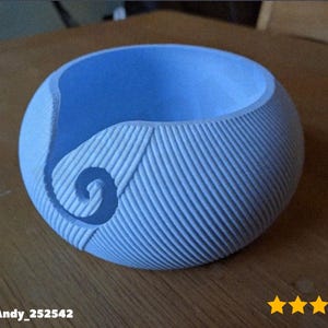 May include: A light blue, spherical yarn bowl with a textured, ribbed exterior. A spiral design is carved into the side. The bowl sits on a wooden surface. The bowl is designed to hold yarn while knitting or crocheting.