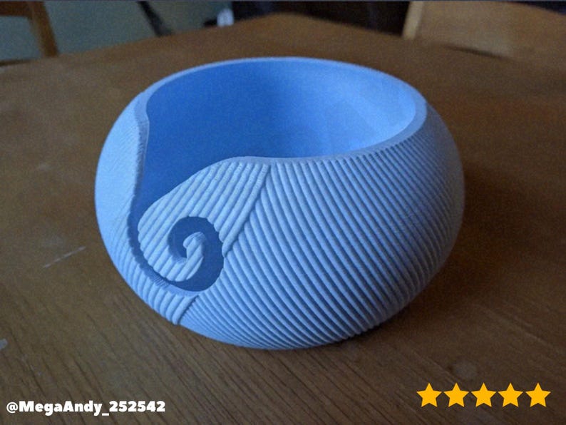 May include: A light blue, spherical yarn bowl with a textured, ribbed exterior. A spiral design is carved into the side. The bowl sits on a wooden surface. The bowl is designed to hold yarn while knitting or crocheting.