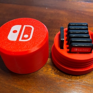 Puede incluir: Caja de plástico roja con un logotipo de Nintendo Switch en la tapa. La caja está abierta para revelar un compartimento para guardar cartuchos de juegos. El compartimento está lleno de cartuchos de juegos de Nintendo Switch negros.