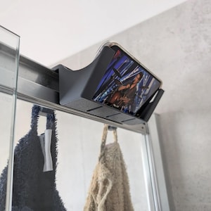 Può includere: Un porta cellulare in plastica nera montato su una porta doccia. Il supporto ha una fessura per un telefono e un bordo rialzato per evitare che il telefono scivoli. Due asciugamani sono appesi alla porta della doccia.