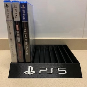 May include: A black plastic stand for video game discs. The stand has slots for multiple discs and features the PlayStation logo and the text "PS5". Three video game cases are displayed on the stand, including "The Last of Us Part II", "Marvel's Spider-Man: Miles Morales", and "Ghost of Tsushima".
