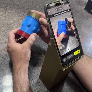 May include: A blue and red cylindrical object with rounded protrusions, held near a smartphone in a gold stand. The phone displays the object and the text "Recorded with my new stand". The stand is angled, and the background is a speckled surface.