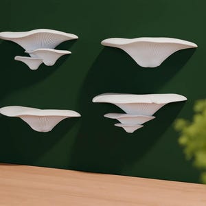 Puede incluir: Cuatro estantes de pared blancos con forma de hongo están montados en una pared verde oscuro. Los estantes varían en tamaño y están dispuestos en un patrón visualmente atractivo. Un jarrón gris con vegetación se encuentra sobre una superficie de madera en primer plano.