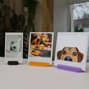 May include: Three photo stands with different colored bases. The stands are holding photos of a camera, a 3D printed object, and a dog. The stands are made of plastic and are about 3 inches tall.