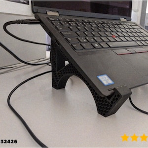 Puede incluir: Ordenador portátil negro sobre un soporte inclinado negro con diseño de panal. El soporte eleva el portátil. Un cable negro está conectado al portátil. La imagen incluye el texto "@finke_132426" y cinco estrellas doradas.