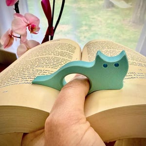 May include: A light teal cat-shaped book page holder. The cat has two small blue eyes and is placed on an open book. The book is open to a page with printed text. Pink orchids are in the background.