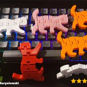 Op de afbeelding: Een verzameling kleurrijke, beweegbare kattenspeeltjes in wit, roze, oranje, groen, rood en grijs. De speeltjes zijn gerangschikt op een zwart toetsenbord. De afbeelding bevat de tekst "@JacekMaryniowski" en een vijfsterrenbeoordeling.