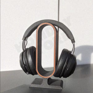 May include: Black over-ear headphones resting on a gray headphone stand with an orange interior. The stand is rectangular with a curved top to hold the headphones. The headphones and stand are on a dark surface.