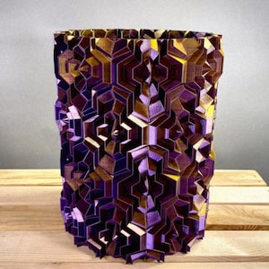May include: A geometric, cylindrical vase with a complex, multi-faceted design. The vase features a combination of purple, gold, and brown tones, creating a striking visual effect. The design is intricate and appears to be 3D printed. The base is resting on a wooden surface.