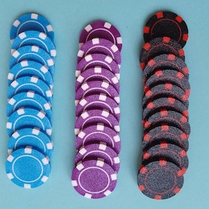 May include: Three stacks of poker chips in blue, purple, and black. The blue chips have white accents, the purple chips have white accents, and the black chips have red accents. The chips are arranged vertically on a light blue surface.