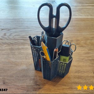 May include: A black desk tidy with several sections, containing scissors, pens, pencils, and a highlighter. The organiser is made of a dark, lattice-like material and is on a wooden surface.