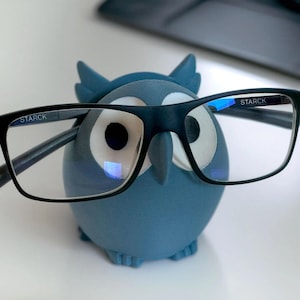May include: A blue owl-shaped glasses holder with the word "STARCK" printed on the glasses.