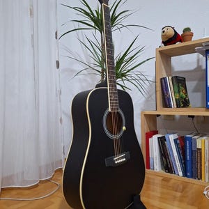 May include: A black acoustic guitar with a wooden stand. The guitar has a black body and a natural wood neck. The guitar is in a room with a plant and a bookshelf.