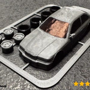 May include: A gray 3D-printed model car kit with a silver body and a brown windshield. The kit includes wheels and tires, all attached to a gray plastic frame. The image has a textured appearance.