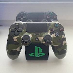 Puede incluir: Un mando de PlayStation 4 de color verde camuflaje con un soporte negro que presenta el logotipo de PlayStation en verde. El mando tiene una placa frontal negra con los botones estándar de PlayStation.