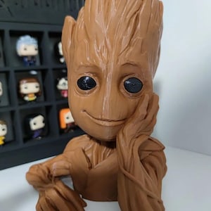 May include: A brown 3D printed figurine of Groot from Guardians of the Galaxy. Groot is sitting with his arm resting on his chin and has a slight smile on his face. His eyes are black and his skin is textured.