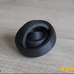 Könnte beinhalten: Ein schwarzes, abstraktes, 3D-gedrucktes Objekt mit einem Spiraldesign. Das Objekt besteht aus einem strukturierten Material und liegt auf einer hellen Holzoberfläche. Das Design ist eine Reihe von verschachtelten, abgerundeten Formen.