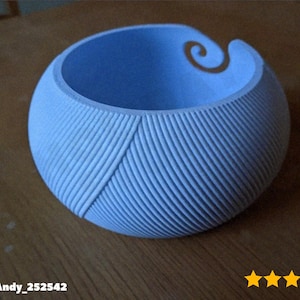 May include: A light blue yarn bowl with a spiral design on the rim. The bowl has a textured, ribbed exterior and a diagonal pattern. The yarn bowl is sitting on a wooden surface.