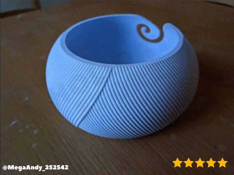 May include: A light blue yarn bowl with a spiral design on the rim. The bowl has a textured, ribbed exterior and a diagonal pattern. The yarn bowl is sitting on a wooden surface.