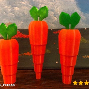 以下が含まれることがあります： 緑色の葉が付いたオレンジ色のニンジン型のオブジェクトが3つ、空と風景が描かれた背景に展示されています。 ニンジンは層状のデザインで、装飾品またはおもちゃである可能性があります。 画像にはEtsyショップ名が含まれています。