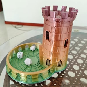 Könnte beinhalten: Ein 3D-gedruckter Würfelturm in Form eines Schlosses. Der Turm ist hauptsächlich kupferfarben mit einem rosa Dach und hat Bogenfenster. Eine grüne und goldene kreisförmige Basis hält mehrere weiße Würfel, ideal für Tabletop-Spiele.