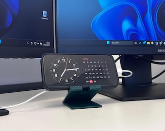 May include: A black smartphone showing a clock and calendar, on a dark green stand. The screen displays the time and the April calendar. The phone is connected to a white charging cable. Two computer monitors are in the background.