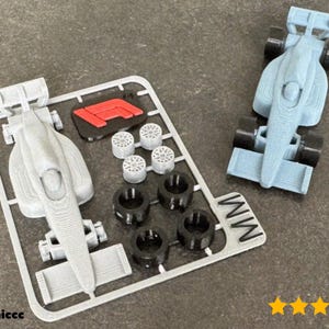 Pode incluir: Um modelo de carro de corrida de Fórmula 1 impresso em 3D em azul claro e cinza, e peças, incluindo rodas e um logotipo vermelho e preto. O modelo está desmontado, com um carro já montado.