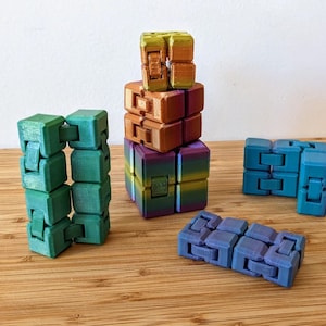 May include: A set of five colorful fidget toys made of plastic. The toys are in various shades of green, blue, purple, orange, and yellow. The toys are all cube-shaped and have a unique design that allows them to be rotated and manipulated.