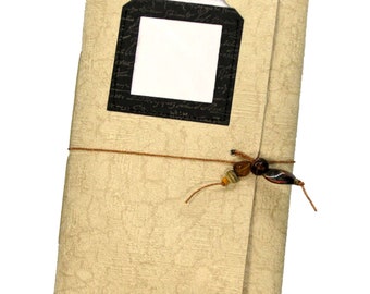 May include: A beige journal with a black frame and a blank white space inside. The journal is tied with a brown string and has brown and orange beads.