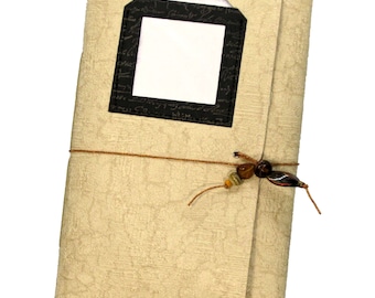 May include: A beige journal with a black frame and a blank white space inside. The journal is tied with a brown string and has brown and orange beads.