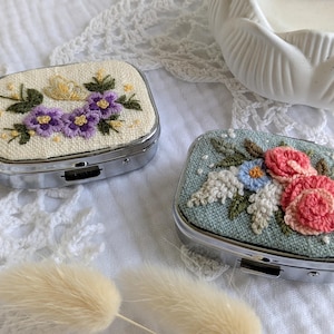 以下が含まれることがあります： 刺繍された花の模様が施された、2つの小さな長方形の金属製ピルケース。片方はクリーム色の背景に紫色の花、もう片方はティール色の背景にピンクと青の花が描かれています。どちらのケースも銀色の金属フレームです。