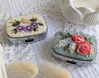 May include: Two small, rectangular metal pill boxes with embroidered floral designs. One box features purple flowers on a cream background, the other has pink and blue flowers on a teal background. Both boxes have a silver metal frame.
