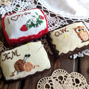 Pode incluir: Três almofadas retangulares com desenhos bordados. Uma tem "I love you" com um coração e morangos. Outra tem uma coruja e as iniciais "O.W.". A terceira tem as iniciais "K.M." e fios. Todas têm bordas decorativas.