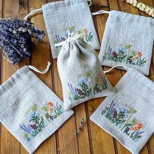 以下が含まれることがあります： 青、オレンジ、緑の色合いで刺繍された野花が特徴の、リネンの巾着袋のコレクション。木製の表面に配置され、乾燥したラベンダーの束が近くにあります。バッグは自然で素朴な美学を持っています。