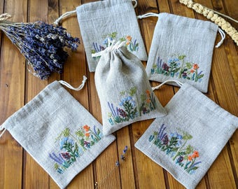 May include: A collection of linen drawstring bags, each adorned with embroidered wildflowers in shades of blue, orange, and green. The bags are arranged on a wooden surface, with a bundle of dried lavender nearby. The bags have a natural, rustic aesthetic.