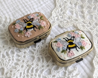 May include: Two rectangular metal pill cases with hinged lids. One case features a tan fabric top with an embroidered bumblebee surrounded by pink and blue flowers. The other case has a white fabric top with an identical bee and floral design.